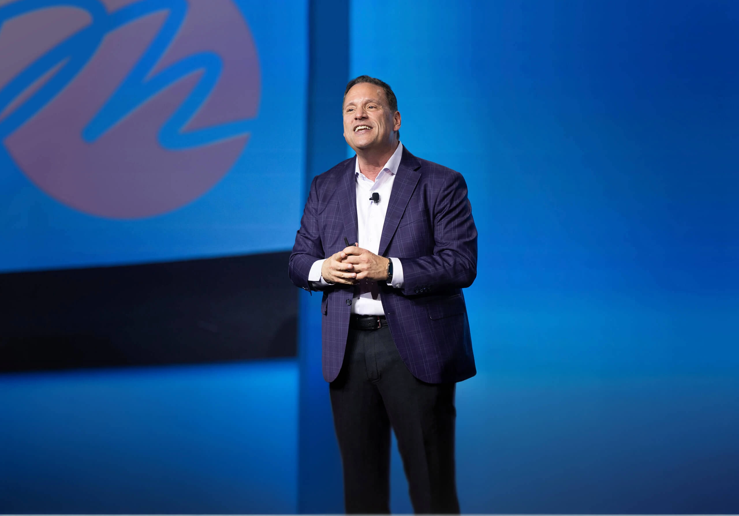 Mark C. Perna speaking on stage at an event in a purple blazer against a blue backdrop